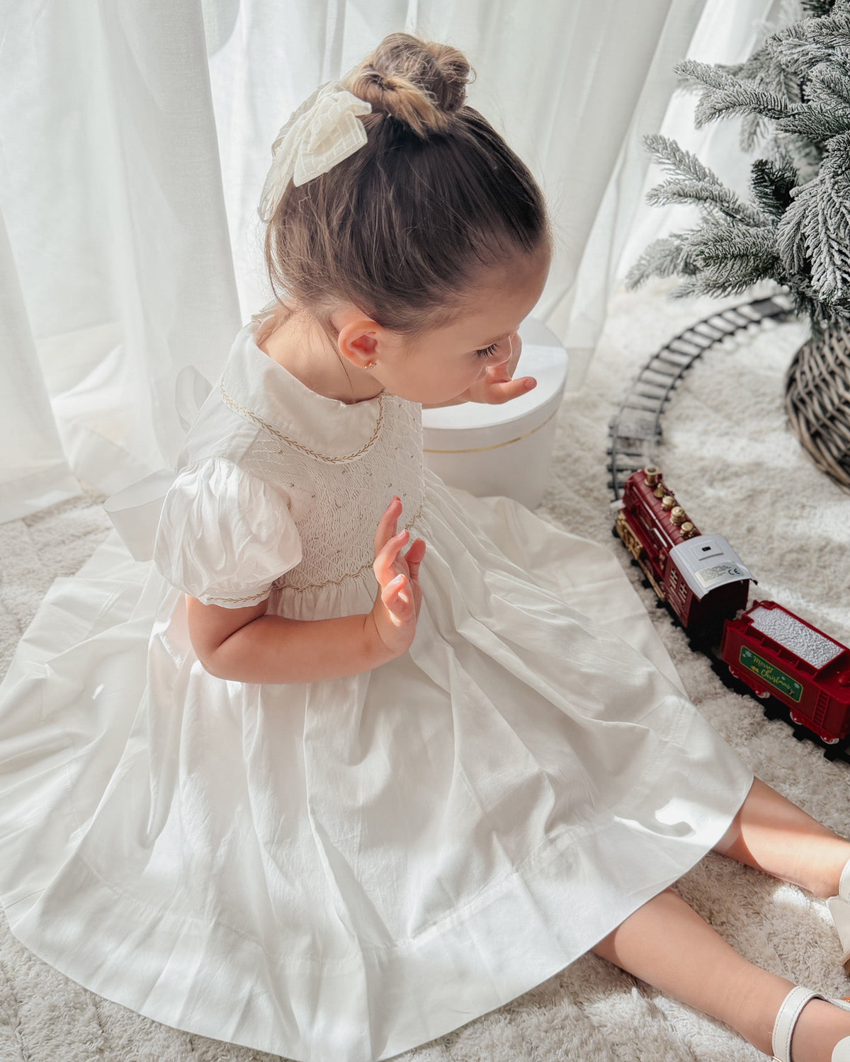 Girl wearing off-white hand smocked dress seated beside a Christmas tree