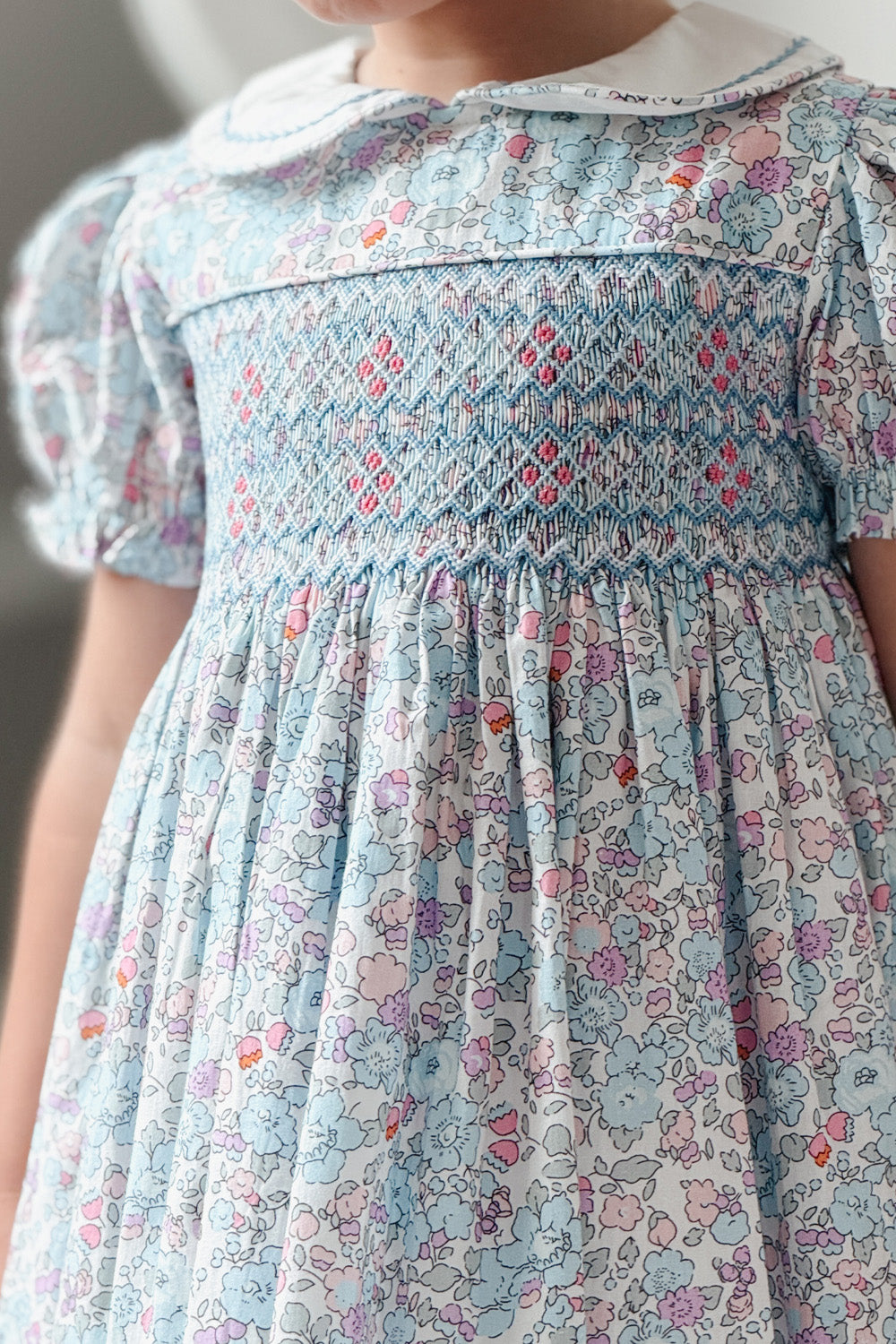 Girl wearing azure blue floral hand smocked dress with Peter Pan collar and puff sleeves