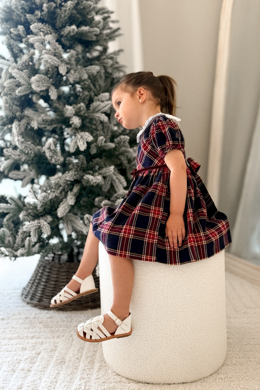 Girl in red and navy tartan dress seated beside a Christmas tree,